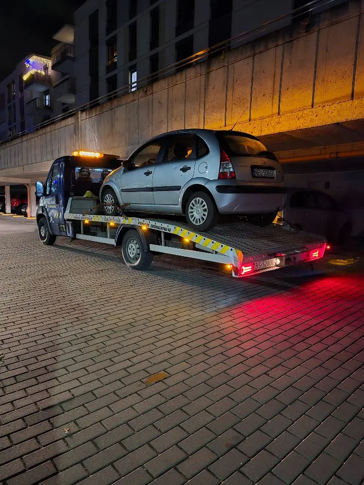 Citroën C3 załadowany na platformę lawety nocą, pod wiaduktem w oświetlonej części miasta.